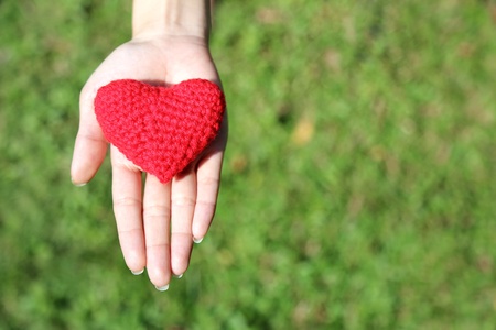Woman hand giving red hand-made crocheted heart with green grass background and copy space. Valentine's Day. Symbol of love.の写真素材