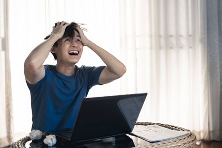 Angry crazy man hand holding head and yelled madly and crumpling paper on table with work on laptop computer in the room, Work from home concept.の写真素材