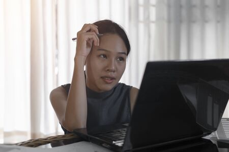 Asian business woman stressed with graph documents and looking at computer screen and hand holding pen and laptop computer on table, Work at Home.の写真素材