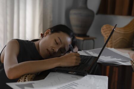 Asian business woman stressed and sleeping on laptop computer with graph documents on table, Work at Home.の写真素材