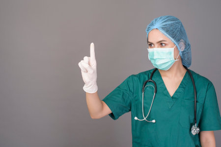 a young confident woman doctor in green scrubs is wearing surgical mask over grey background studioの写真素材