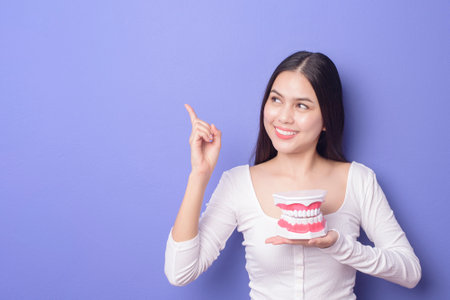 a Young beautiful smiling woman is holding plastic denture teeth over isolated purple background...