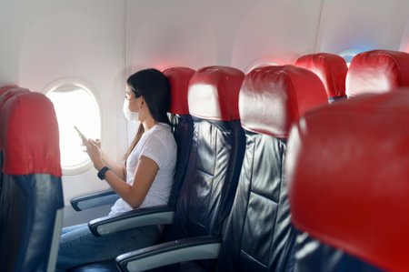A traveling woman wearing protective mask onboard in the aircraft using smartphone, travel under Covid-19 pandemic, safety travels, social distancing protocol, New normal travel conceptの写真素材