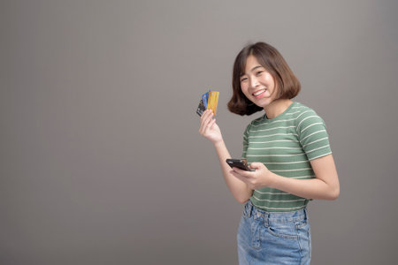 A portrait of young beautiful asian woman holding credit card and smartphone isolated over gray background studioの写真素材