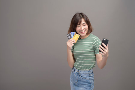 A portrait of young beautiful asian woman holding credit card and smartphone isolated over gray background studioの写真素材