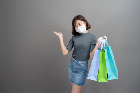 A portrait of young beautiful asian woman wearing a surgical mak is holding credit card and colorful shopping bag isolated over gray background studioの写真素材