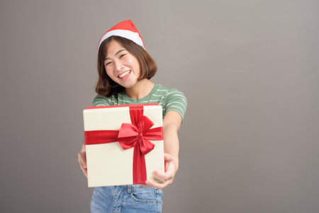 A portrait of beautiful woman wearing red Santa Claus hat holding gift box over studio background,...