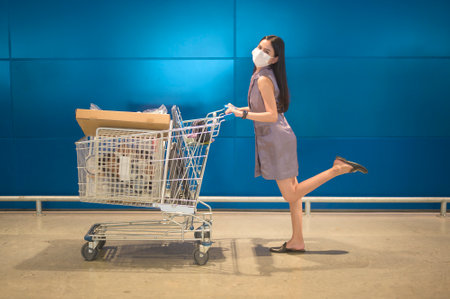 A young woman wearing a surgical mask with a trolley in shopping mall,     pandemic concept.の写真素材