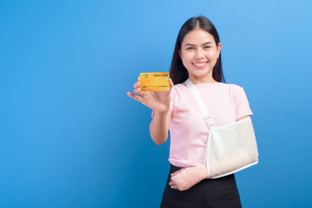 A portrait of young woman with an injured arm in a sling holding a credit card or medical insurance card over blue background in studio, insurance and healthcare conceptの写真素材