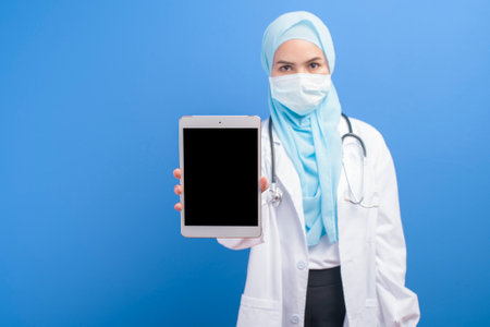 A female muslim doctor with hijab wearing a surgical mask using tablet over blue background studio.の写真素材