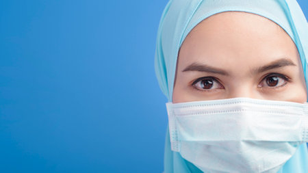A female muslim doctor with hijab wearing a surgical mask over blue background studio.の写真素材