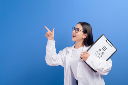 A young female ophthalmologist with glasses holding eye chart over blue background studio, healthcare conceptの写真素材
