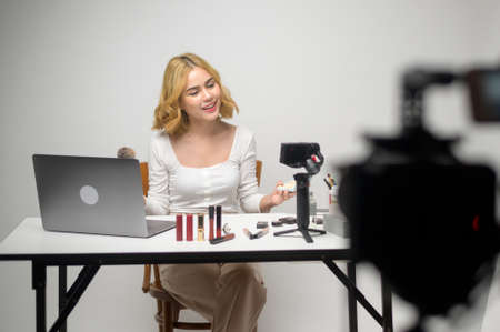 Behind the scenes young blonde woman entrepreneur working with laptop presents cosmetic products during online live stream over white background studio, selling online and beauty blogger conceptの写真素材