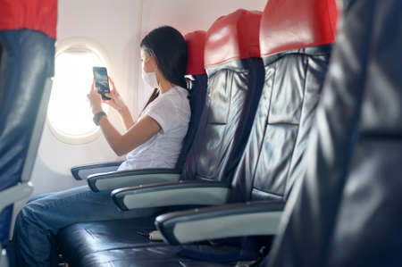 A traveling woman wearing protective mask onboard in the aircraft using smartphone, travel under Covid-19 pandemic, safety travels, social distancing protocol, New normal travel conceptの写真素材