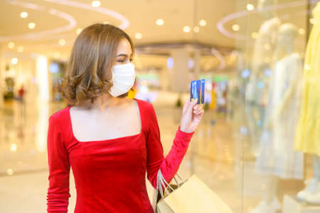A young woman in red dress wearing protective mask in shopping mall, shopping under pandemic...
