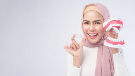 A young muslim woman holding invisalign braces and an artificial Dental Model Of Human over white...