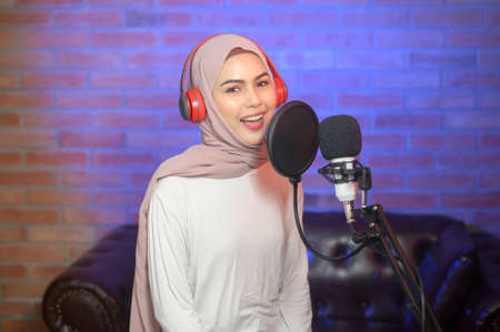 A young smiling muslim female singer wearing headphones with a microphone while recording song in a music studio with colorful lights.の写真素材