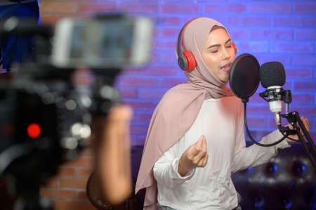 A young smiling muslim female singer wearing headphones with a microphone while recording song in a music studio with colorful lights.の写真素材