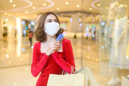 A young woman in red dress wearing protective mask in shopping mall, shopping under Covid-19 pandemic concept.の写真素材