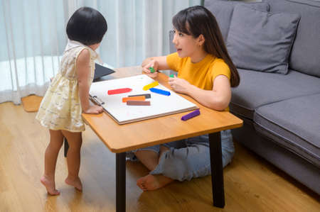 A young mom helping daughter drawing with colored pencils in living room at home.