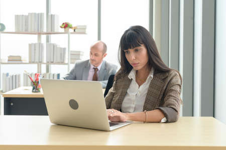 A young businesswoman sitting and working with laptop at modern office with colleague behindの写真素材