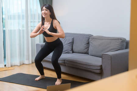 A young woman is exercising and watching online training fitness class on the laptop in living room...