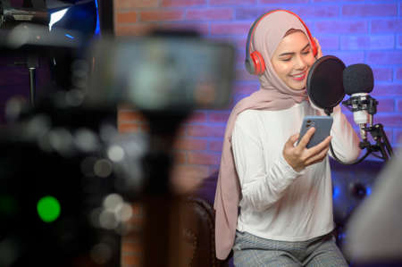 A young smiling muslim female singer wearing headphones with a microphone while recording song in a music studio with colorful lights.の写真素材