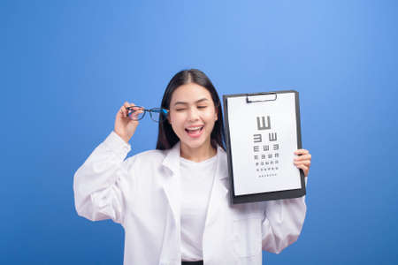 A young female ophthalmologist with glasses holding eye chart over blue background studio, healthcare conceptの写真素材