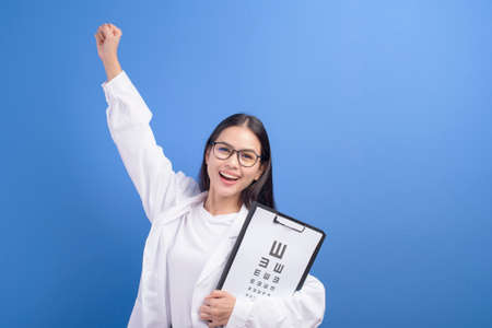 A young female ophthalmologist with glasses holding eye chart over blue background studio, healthcare conceptの写真素材