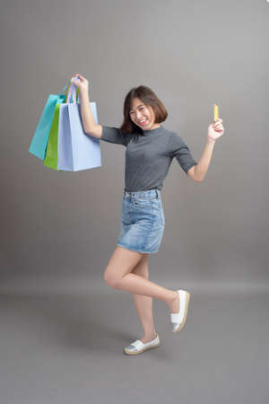A portrait of young beautiful asian woman holding credit card and colorful shopping bag isolated over gray background studioの写真素材