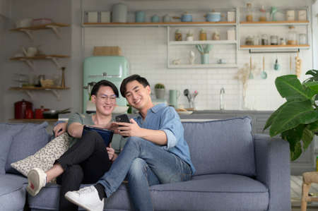 Young smiling gay couple using smartphone and relaxing in the living room at home, LGBTQ and diversity concept.の写真素材