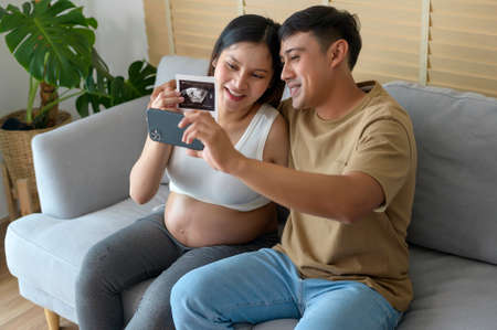 Young pregnant woman with husband embracing and video call with family and friends by smartphone on social media, family and pregnancy care conceptの写真素材