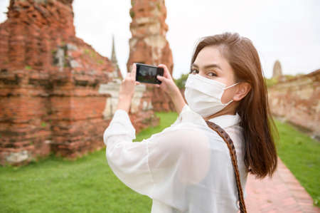 young beautiful woman wearing protective mask traveling and taking photo at thai historical Park, Holidays and cultural tourism concept.の写真素材