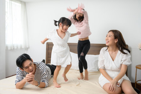 Asian Parents with two little daughters jumping on the bed in bedroom, happy family conceptの写真素材