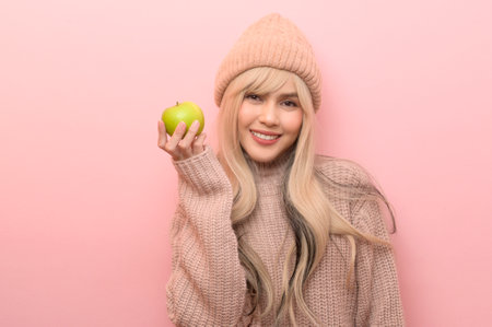 Portrait of Caucasian young woman wearing sweater holding green apple over pink backgroundの写真素材