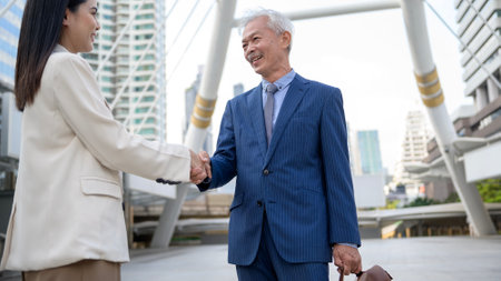 Asian senior mature middle aged businessman handshaking young businesswoman in modern cityの写真素材