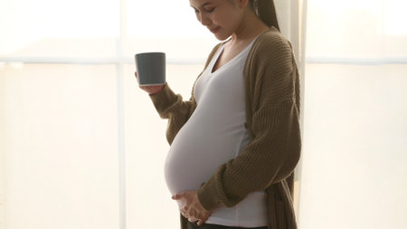A pregnant woman gently cradles her belly while sipping a warm beverage in natural light.の写真素材