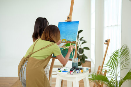 Two young women are painting landscapes on easels in a well-lit art studio.