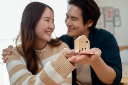Couple smile joyfully while holding a small house model in a bright living space.の写真素材