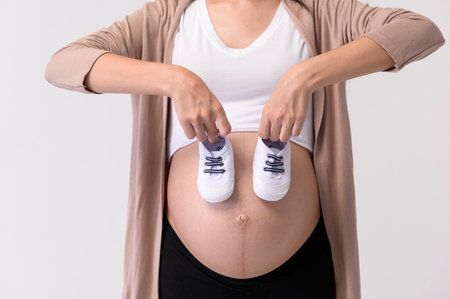Portrait of Beautiful pregnant woman holding baby shoes over white background studio