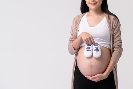 Portrait of Beautiful pregnant woman holding baby shoes over white background studio