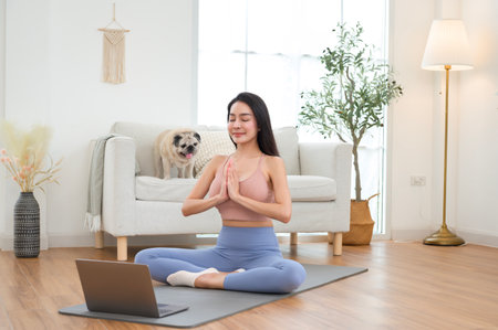 A woman meditates on a yoga mat, streaming a class on her laptop in a cozy living room.の写真素材