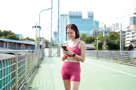 A woman jogs along a city path, smiling as she checks her phone and listens to music.の写真素材