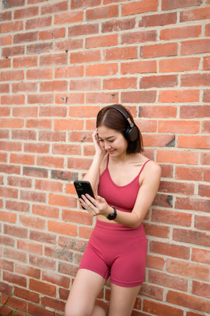 A woman in athletic clothing enjoys music on her smartphone while leaning against a brick wall.の写真素材