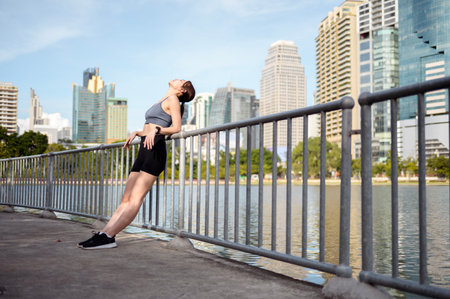 A woman stands by a lake in a city, embracing the warm sun as she enjoys outdoor fitness.