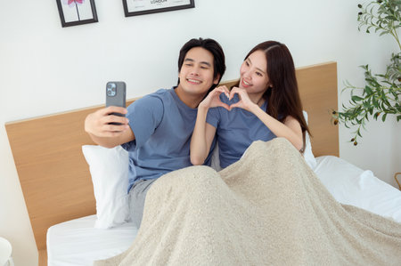 A couple sits on their bed, sharing laughter while looking at a phone together in a bright room.