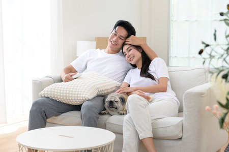 A joyful couple sits together on a sofa, sharing snacks with their cute pug in a sunny space.の写真素材