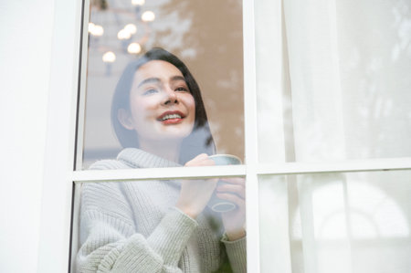 A woman in a sweater sips a warm drink, looking content while standing by the window.