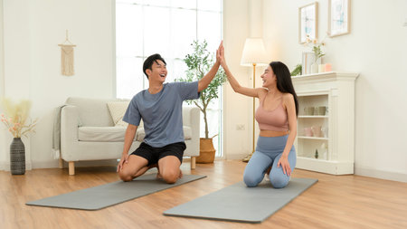 Two individuals perform yoga poses in a bright room with fitness mats and comfortable seating.