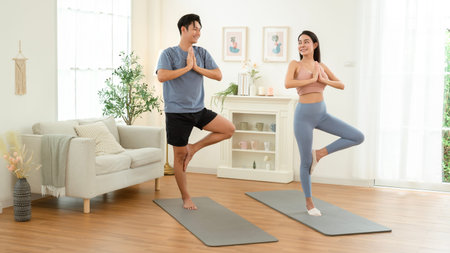 Two individuals perform yoga poses in a bright room with fitness mats and comfortable seating.